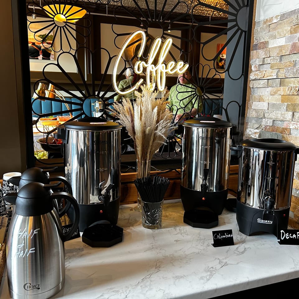 A coffee station with three large dispensers, a "Coffee" neon sign, and a half-and-half carafe.