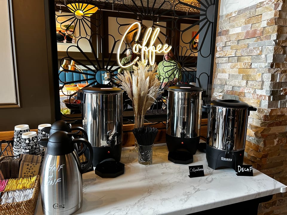 A coffee station with three large dispensers, a "Coffee" neon sign, and a half-and-half carafe.